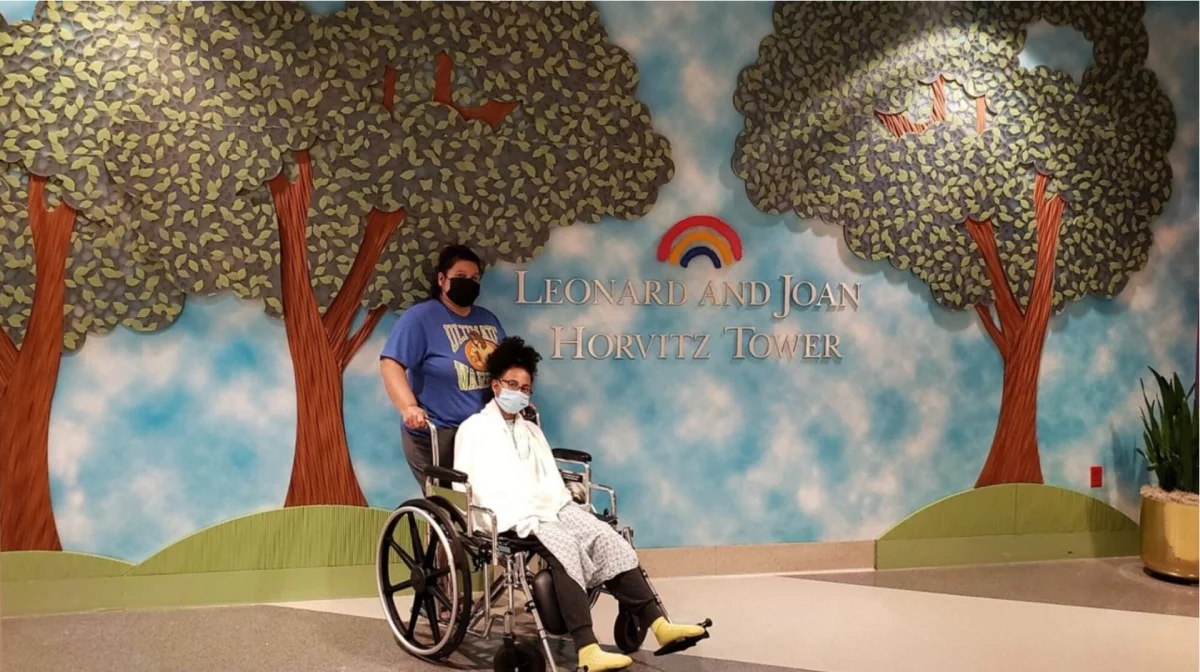 Una persona en silla de ruedas y otra de pie a su lado posan delante de un mural con árboles y el texto "Torre Leonard y Joan Horvitz". Ambos llevan máscaras.