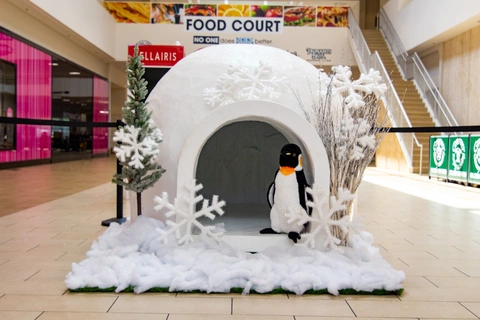 Un centro comercial con un iglú rodeado de nieve artificial y árboles blancos. Un pingüino de peluche en la entrada. Ambiente luminoso y festivo.
