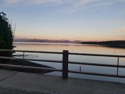 Tranquilo lago al atardecer con montañas a lo lejos, parcialmente enmarcado por la barandilla de un puente. El cielo está suavemente coloreado, creando una atmósfera serena.
