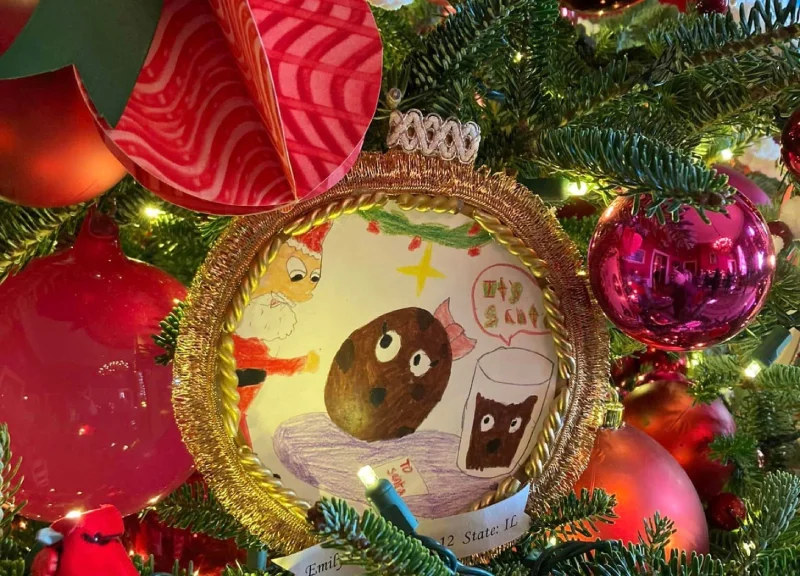 Primer plano de la decoración de un árbol de Navidad con el dibujo de un niño enmarcado en dorado. El colorido dibujo representa galletas animadas y leche.