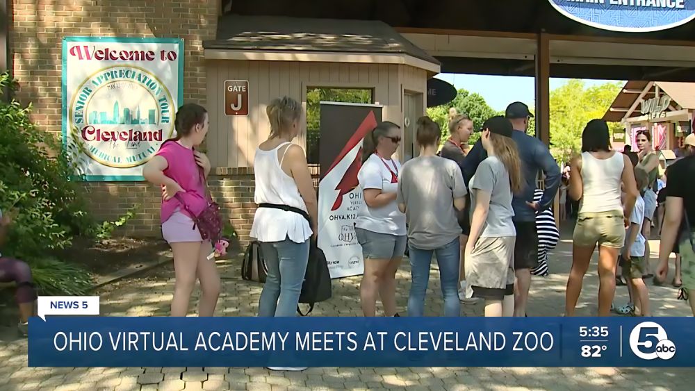 Un grupo de personas se reúne cerca de una puerta del zoo de Cleveland. Se ve un cartel de "Bienvenidos a Cleveland". El ambiente es informal y animado.