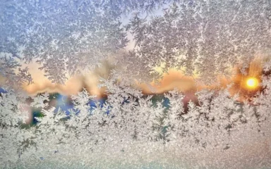 Ventana esmerilada con intrincados dibujos de hielo, que revela un suave y colorido amanecer en el fondo. La escena transmite una mañana serena y fría.