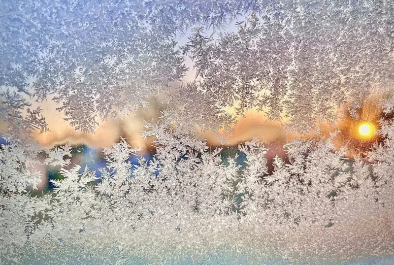 Ventana esmerilada con intrincados dibujos de hielo, que revela un suave y colorido amanecer en el fondo. La escena transmite una mañana serena y fría.