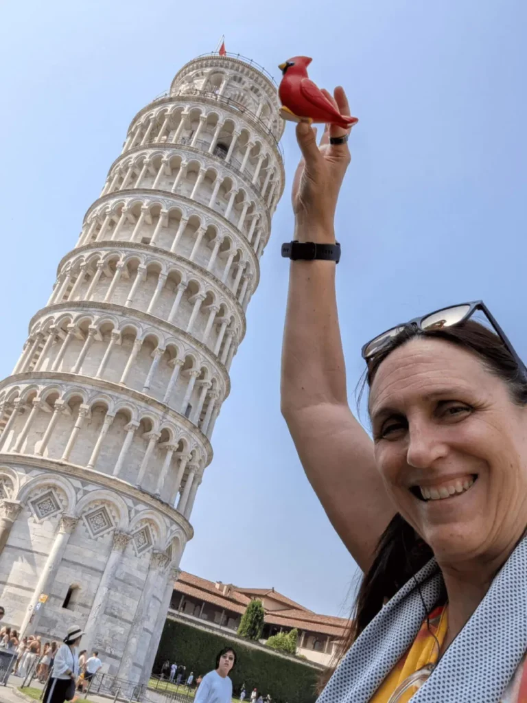 Una mujer sonríe y sostiene una figurita de un cardenal rojo que parece descansar sobre la Torre Inclinada de Pisa, al fondo. La torre se inclina notablemente contra el cielo azul claro, con otros turistas visibles en la base.