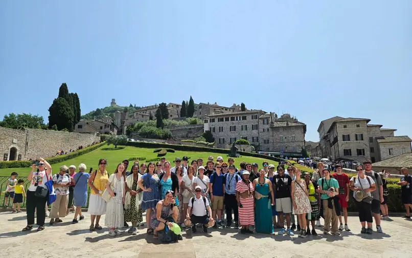 Un gran grupo de viajeros posan juntos al aire libre frente a un pueblo en la ladera de una colina con edificios de piedra, verdes prados y altos cipreses bajo un cielo azul despejado. El grupo está muy unido, sonriendo, y algunos llevan sombreros, gafas de sol o mochilas.