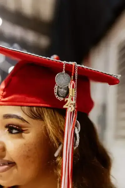 Un graduado con una gorra roja con la borla "23" sonríe suavemente. La gorra lleva un colgante con la foto de una persona, creando un tono festivo y sentimental.