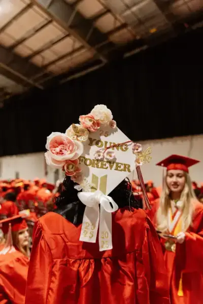 Graduado con toga roja y birrete decorado con flores y "Young Forever". Rodeado de otros graduados, transmitiendo celebración y logro.