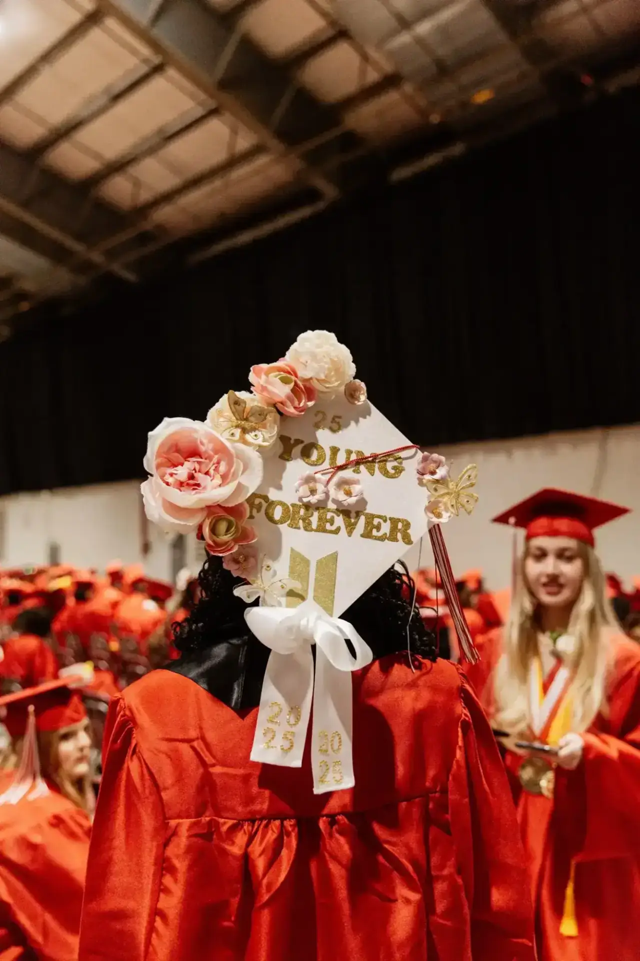 Graduado con toga roja y birrete decorado con flores y "Young Forever". Rodeado de otros graduados, transmitiendo celebración y logro.