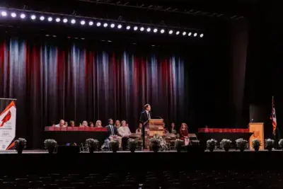 Un hombre se dirige al público desde un podio situado en un gran escenario con telón de fondo. En el escenario hay personas sentadas y plantas en macetas. El ambiente es formal.
