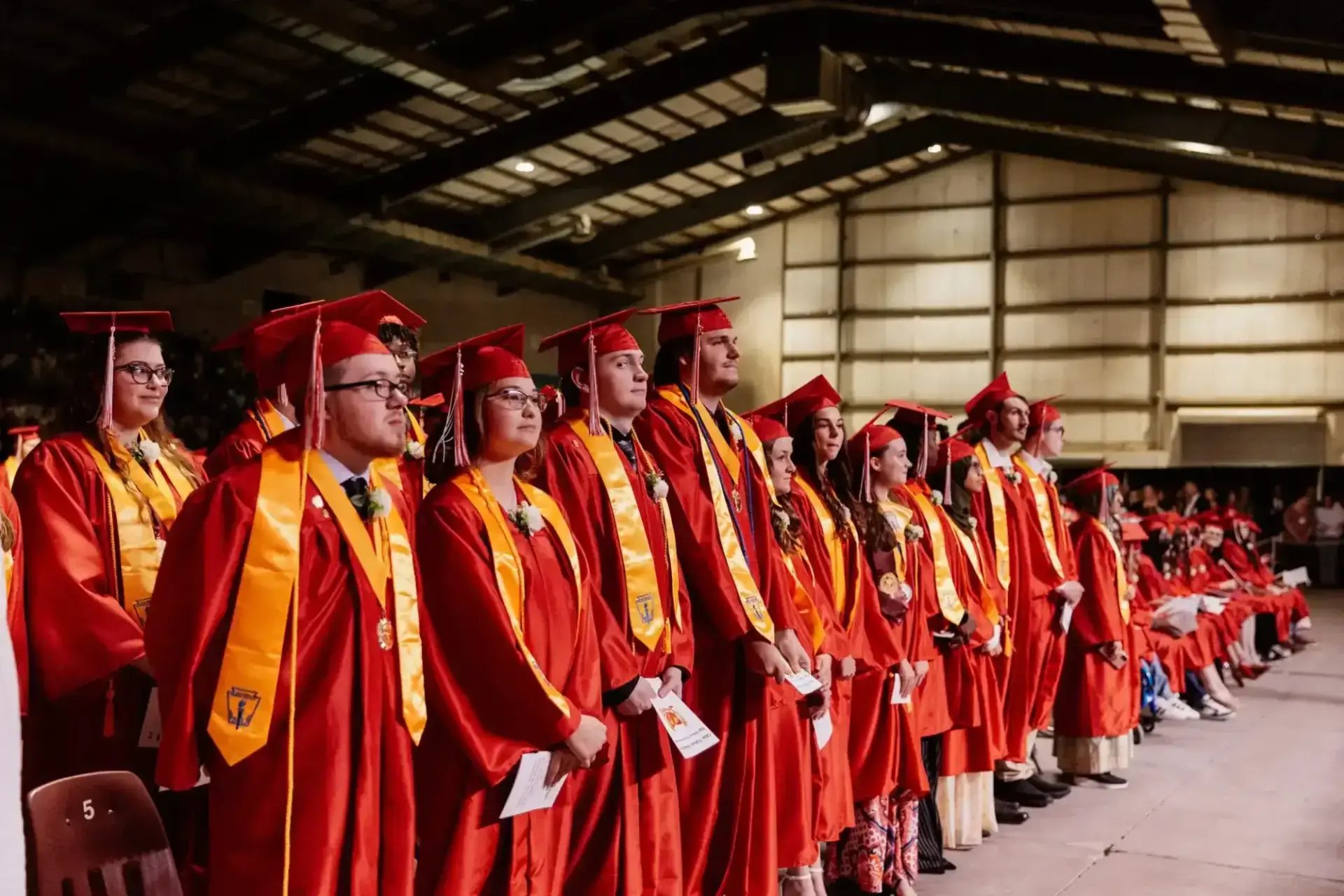 Un grupo de graduados, con togas y birretes rojos, permanecen en fila en el interior, mirando al frente con expectación y orgullo.