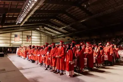 Graduados con togas y birretes rojos se colocan solemnemente en filas durante una ceremonia en una gran sala. Las banderas de EE.UU. y Ohio ocupan un lugar destacado en el fondo.