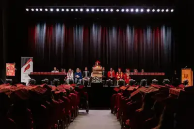 Ceremonia de graduación con estudiantes con togas y birretes rojos sentados frente a un escenario. Un orador en el podio está flanqueado por profesores.