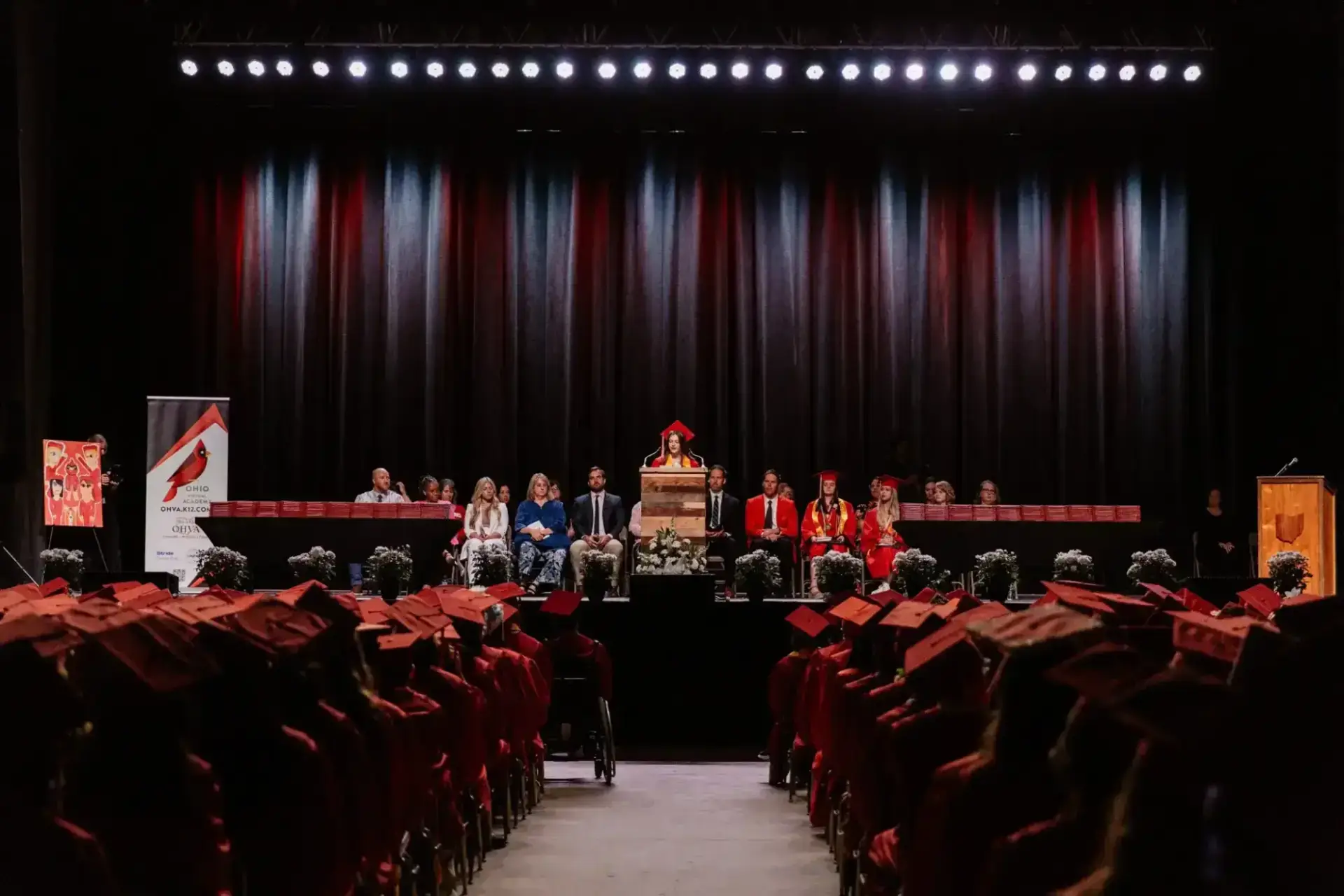 Ceremonia de graduación con estudiantes con togas y birretes rojos sentados frente a un escenario. Un orador en el podio está flanqueado por profesores.