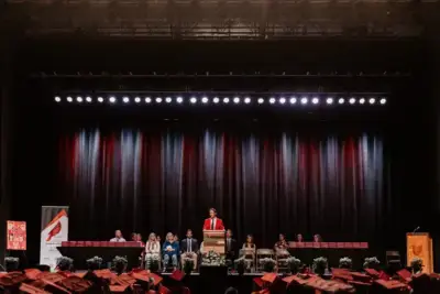 Un orador vestido con una toga roja sube a un podio en un escenario, ante la mirada de un público de graduados con gorras rojas.