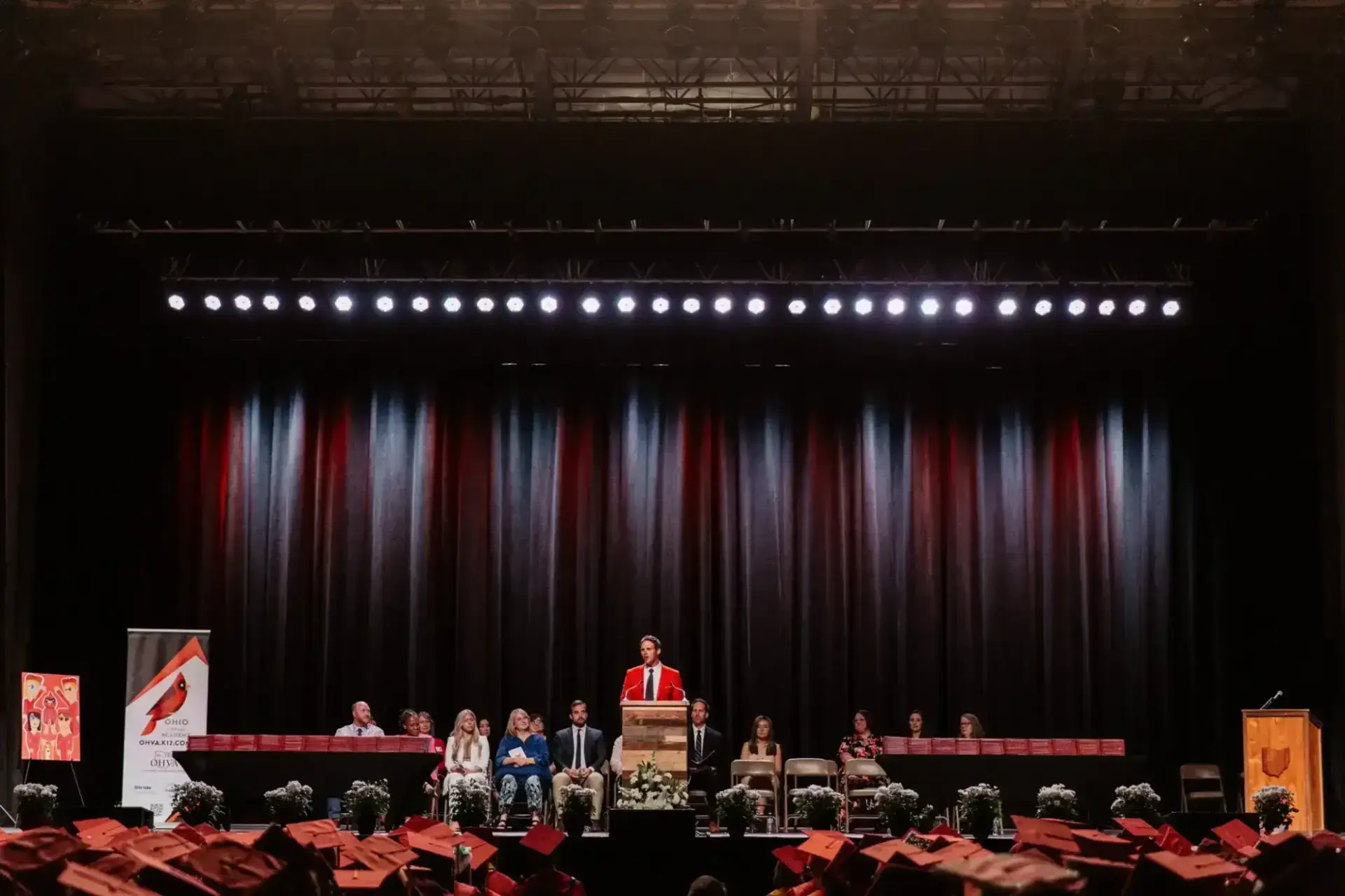 Un orador vestido con una toga roja sube a un podio en un escenario, ante la mirada de un público de graduados con gorras rojas.