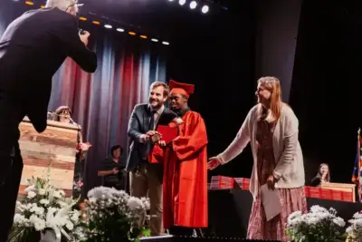 Un graduado con toga y birrete rojos sostiene orgulloso su diploma, flanqueado por dos adultos sonrientes.