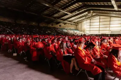 Un gran grupo de estudiantes con togas y birretes rojos se sientan en filas durante una ceremonia de graduación en el interior de un amplio auditorio lleno de espectadores.