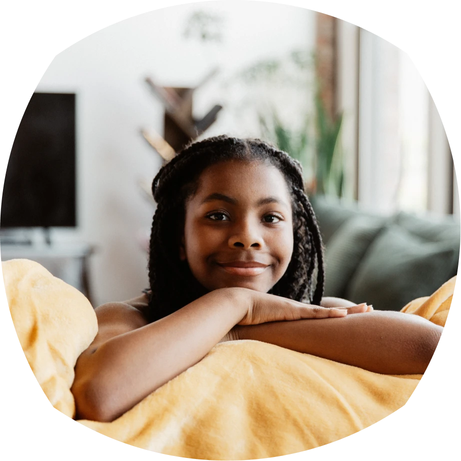 Una joven con el pelo trenzado se apoya en un sofá, sonriendo cálidamente. Está envuelta en una manta amarilla, con un acogedor salón de fondo.
