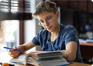 Un joven con gafas estudia en un escritorio, con un bolígrafo en la mano y leyendo un libro. El entorno es tranquilo y concentrado, con luz natural procedente de una ventana.
