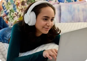 Una joven con auriculares sonríe mientras utiliza un ordenador portátil, tumbada en una cama con almohadas de colores. Parece ocupada y relajada en un entorno acogedor.