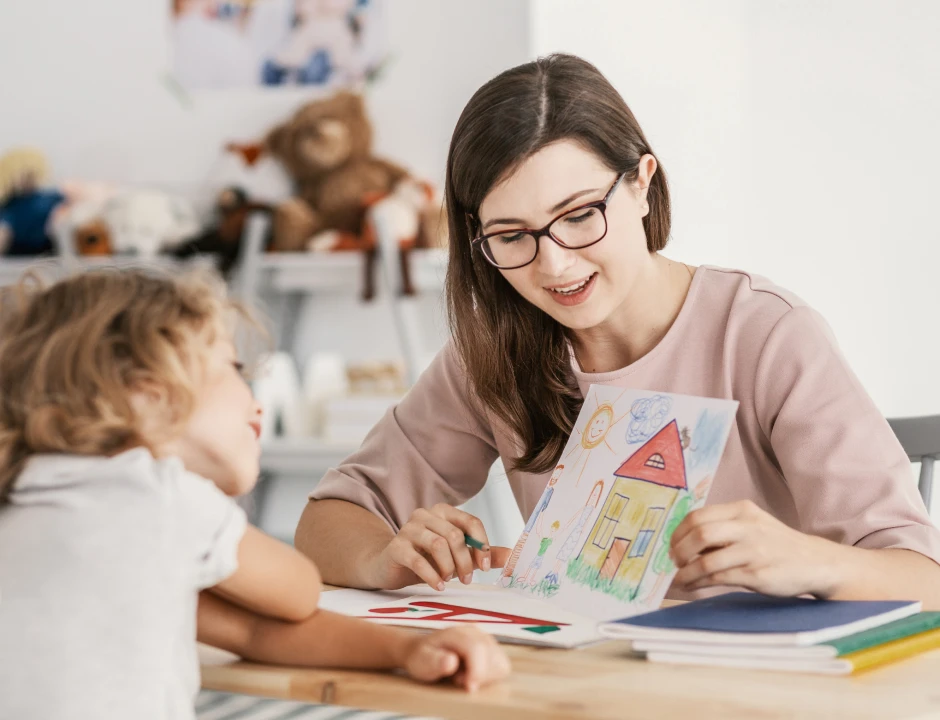 Una mujer sonriente con gafas muestra un colorido dibujo de una casa a un niño pequeño que observa atentamente. La escena, cálida y acogedora, está ambientada en una sala de juegos con juguetes.