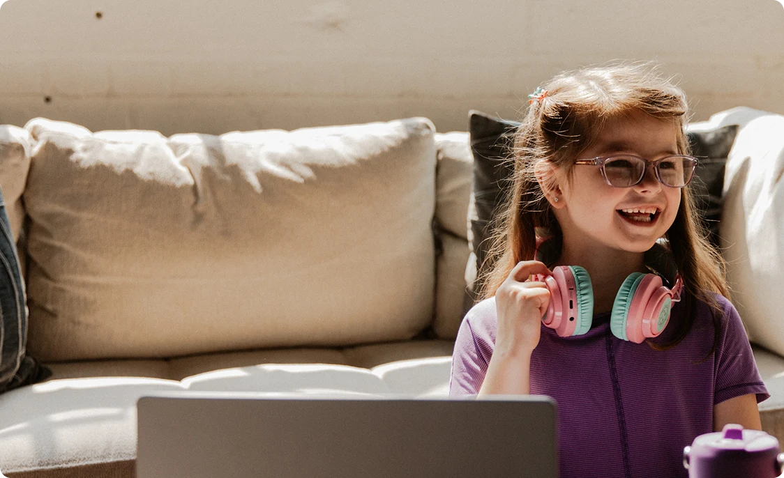 Una joven sonriente con gafas y auriculares de colores se sienta frente a un ordenador portátil en una habitación iluminada por el sol, desprendiendo felicidad y una sensación de confort.