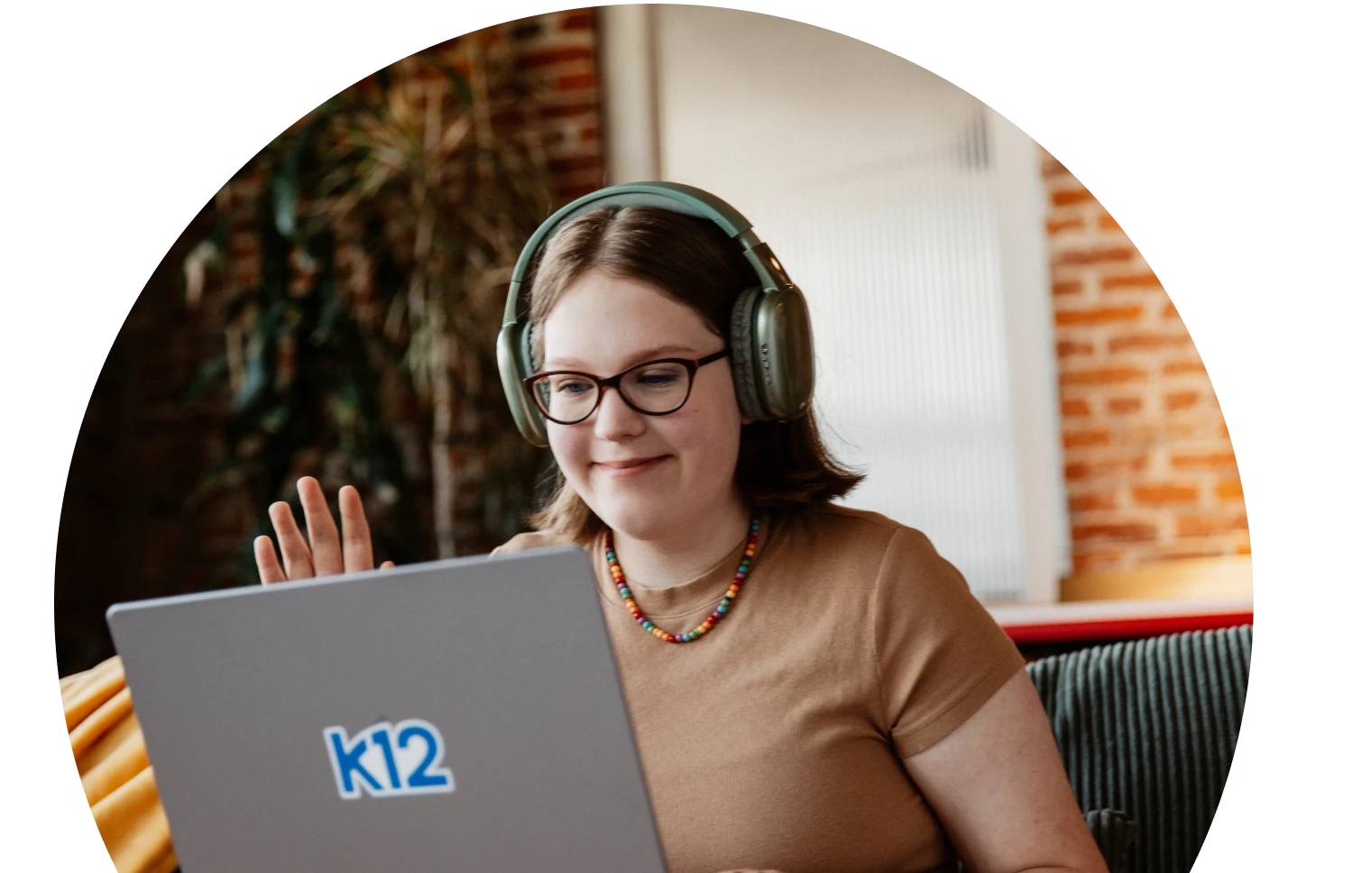 Mujer joven con auriculares y gafas sonríe mientras saluda a un ordenador portátil. Lleva un collar de colores. El fondo muestra una pared de ladrillo y plantas.