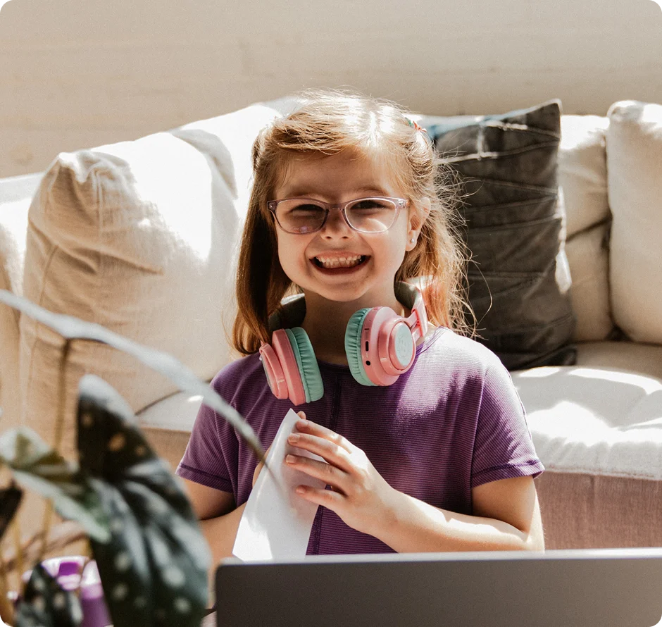 Una joven sonriente con gafas y auriculares rosas está sentada en un sofá, sosteniendo un periódico. La luz natural y un ambiente acogedor llenan la habitación.