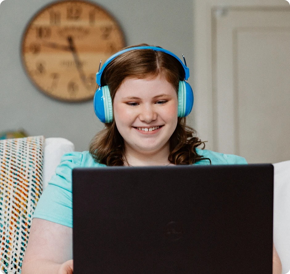 Chica sonriente con auriculares azules utiliza un ordenador portátil en una habitación acogedora. Un reloj de pared redondo y una manta estampada se ven al fondo.