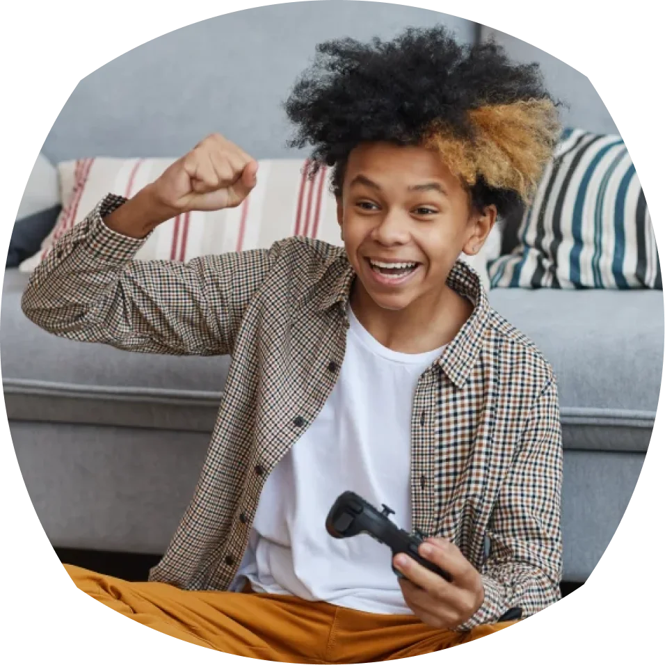 Un joven con camisa de cuadros y pelo rizado sostiene emocionado un mando de videojuegos, puño en alto, sentado en un sofá con cojines a rayas.