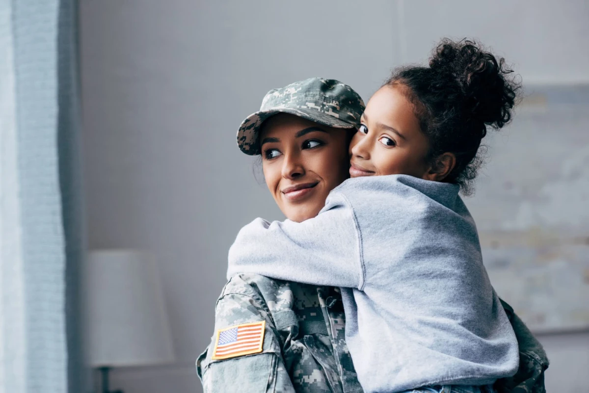 Una mujer con uniforme militar sonríe suavemente mientras una niña la abraza cariñosamente, ambas rebosan felicidad y amor. En el uniforme se ve un parche con la bandera estadounidense.
