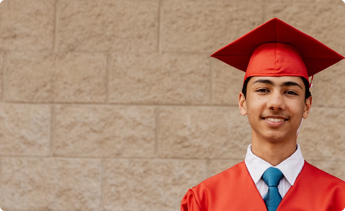Un graduado sonriente con toga y birrete rojos frente a una pared de ladrillo claro. Lleva una camisa blanca y una corbata verde azulado, exudando orgullo y logro.