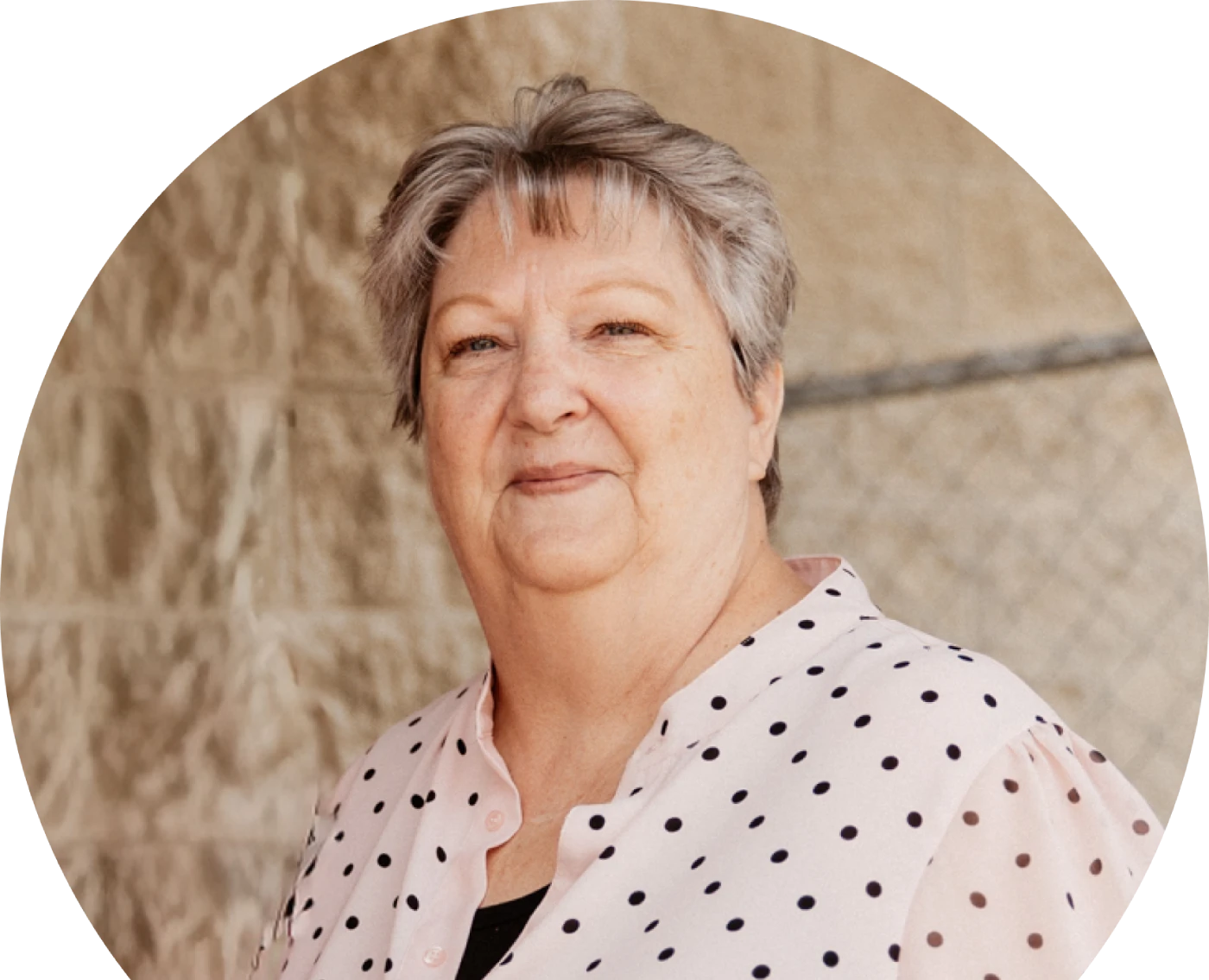 Anciana de pelo corto y canoso sonríe suavemente vestida con una blusa blanca de lunares. El fondo muestra una pared beige con textura y una valla de alambre.