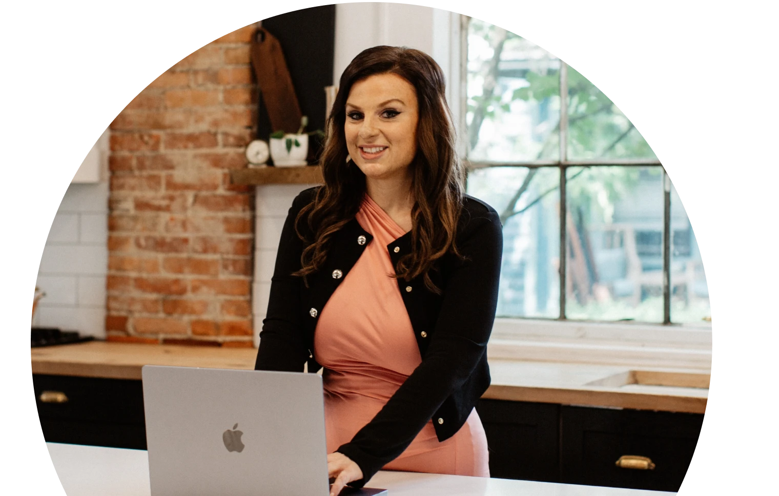 Una mujer con un vestido rosa y una rebeca negra está de pie en la encimera de una cocina con un ordenador portátil, sonriendo con confianza. El fondo tiene paredes de ladrillo y una gran ventana.