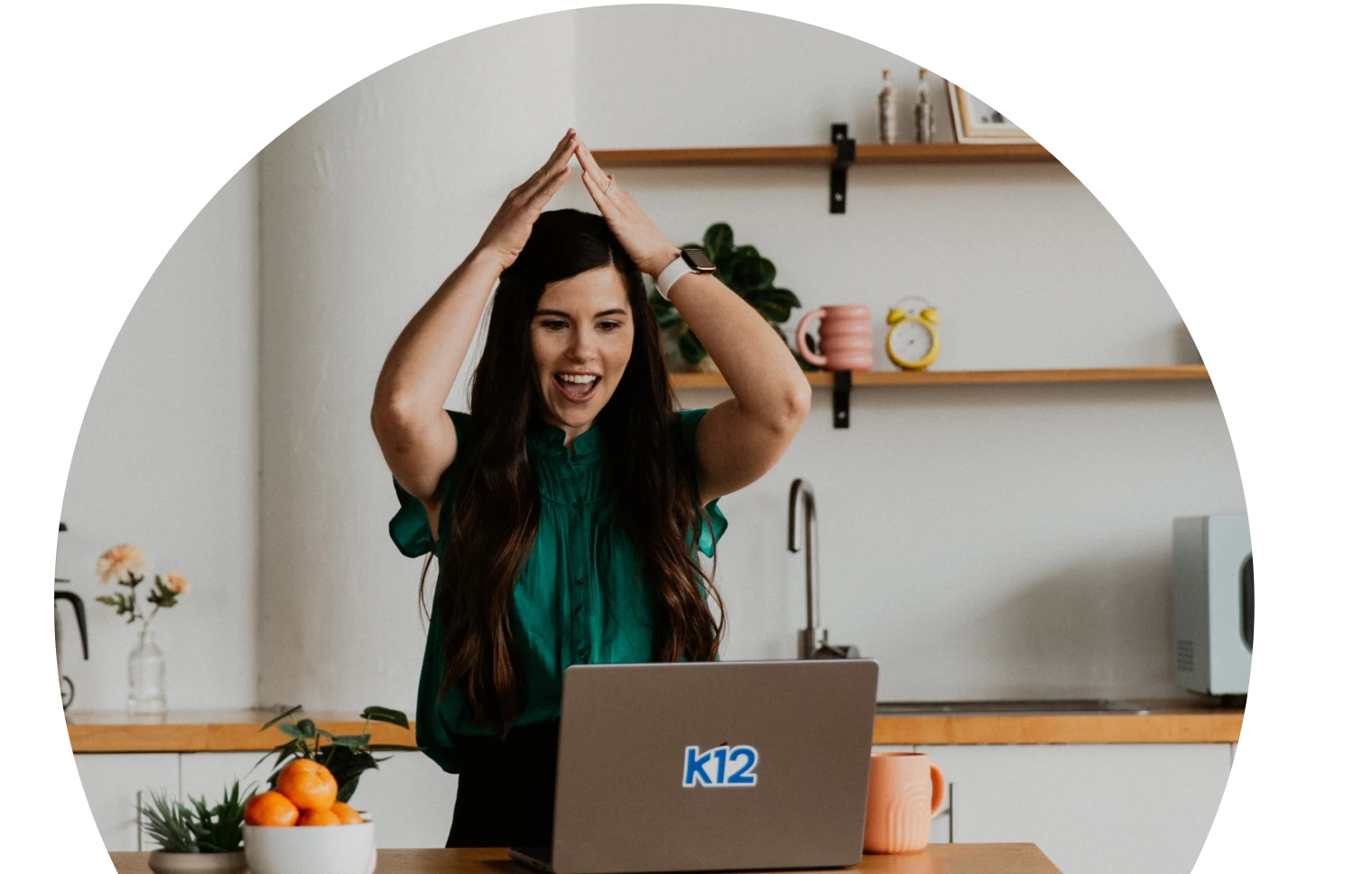 Una mujer con blusa verde azulado gesticula alegremente emocionada delante de un ordenador portátil. El fondo es una acogedora cocina con estanterías, plantas y una decoración vibrante.