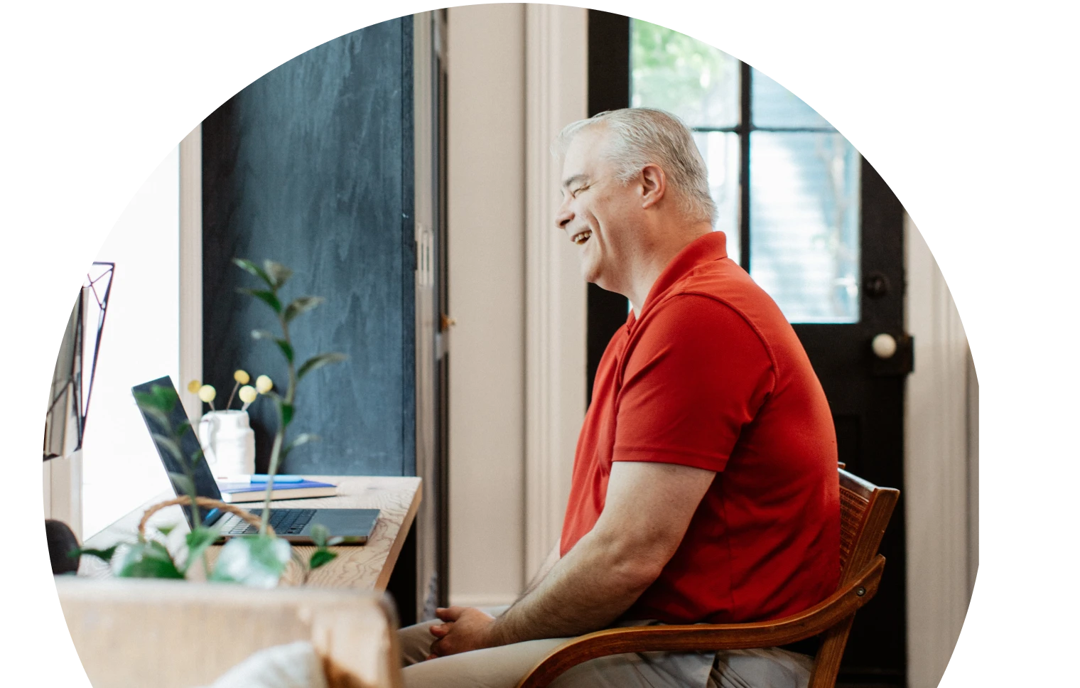 Un hombre con un polo rojo está sentado en un escritorio, sonriendo ante un ordenador portátil. La habitación es luminosa, con una planta y una gran ventana, lo que crea un ambiente relajado.